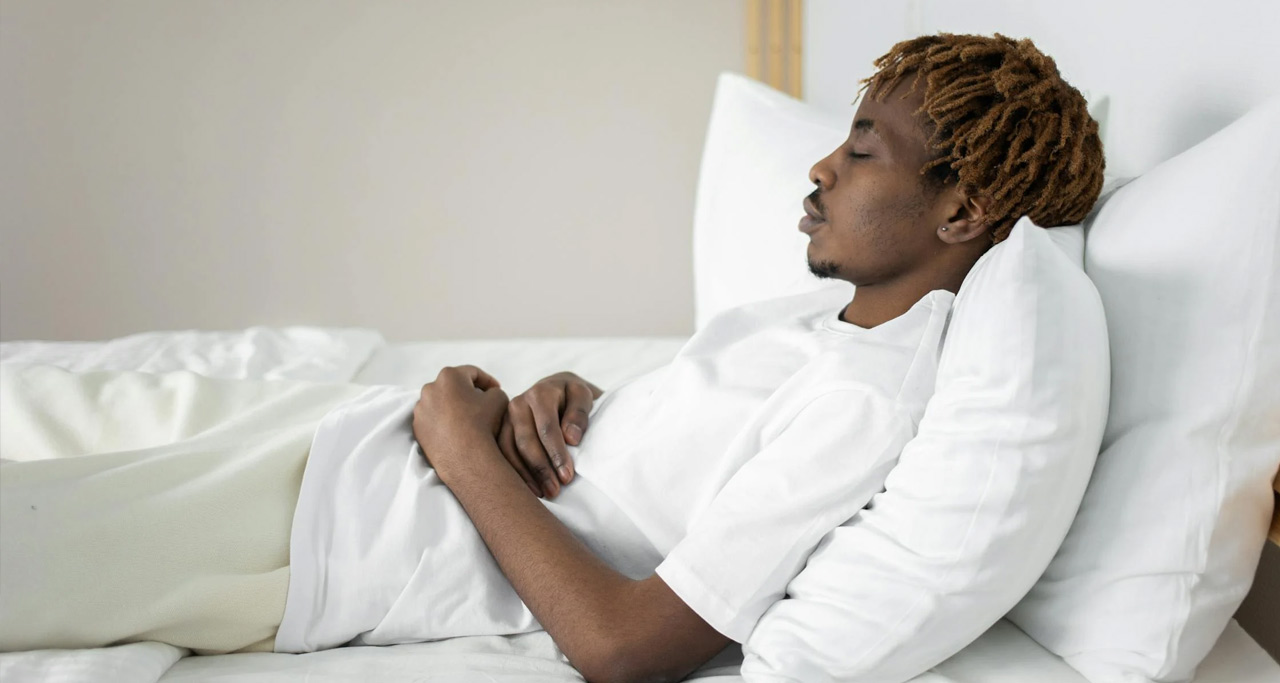 Homem negro deitado em cama com lençóis brancos, vestido de branco.