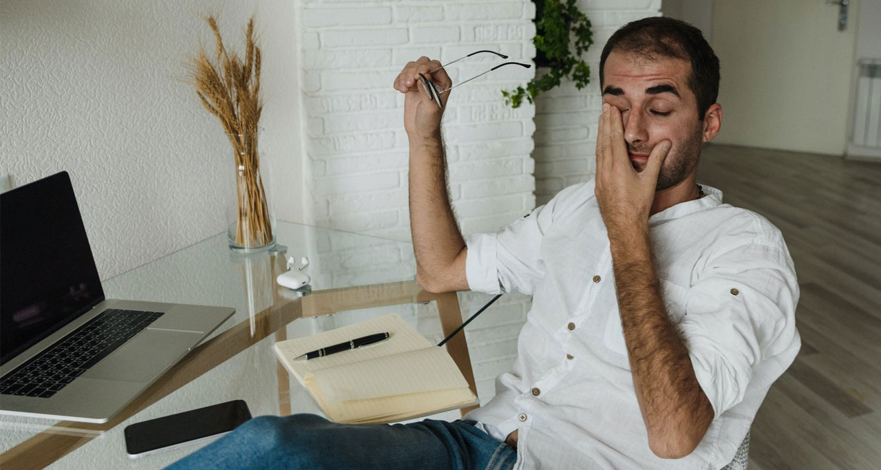 Homem em mesa de trabalho aparentemente cansado, passando a mão nos olhos.