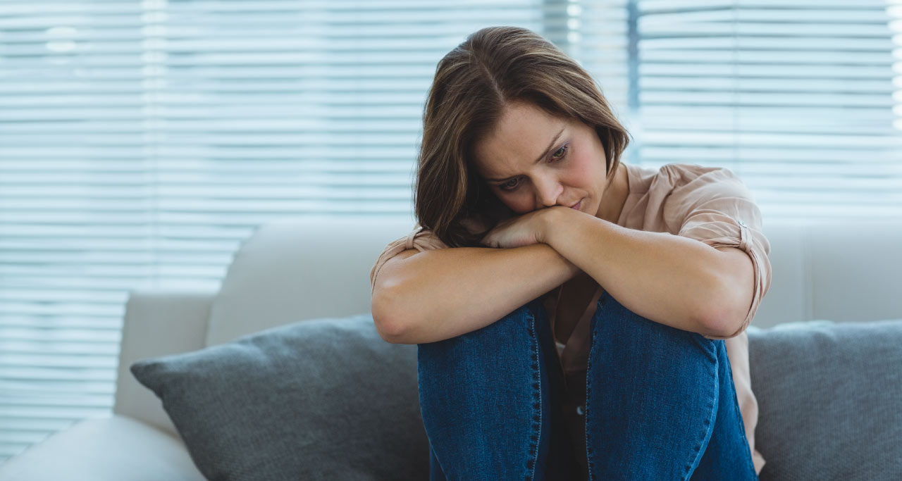 Mulher com depressão sentada no sofá, abraçando seus joelhos.