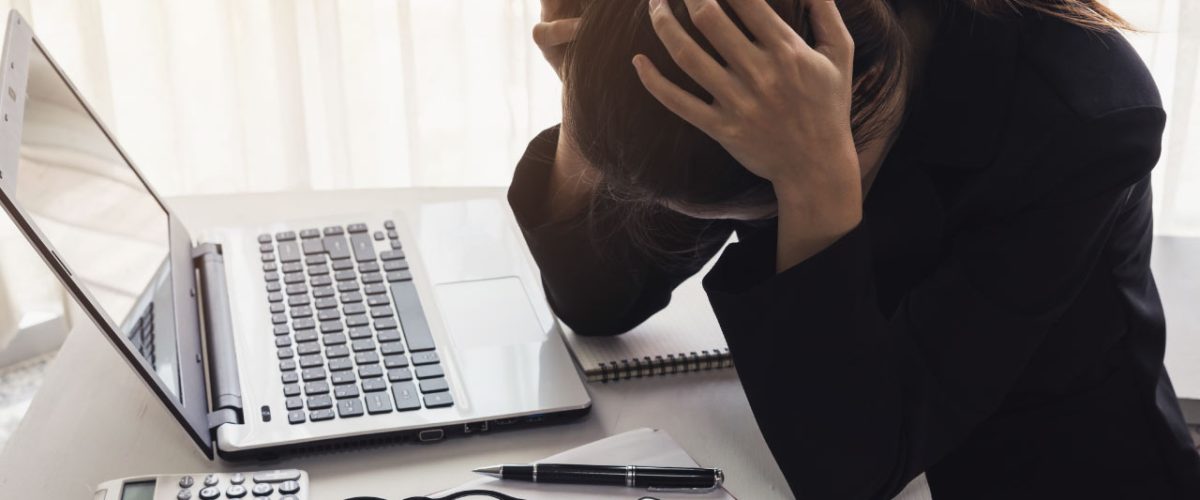 depressao-no-trabalho Mulher segurando a própria cabeça, sobre a mesa de trabalho, demonstrando estresse.