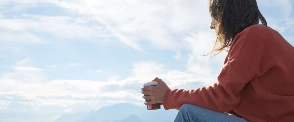 depressão refratária Mulher com calça jeans e casaco rosa segura xícara de café, sentada, pensativa. O fundo é um céu azul.