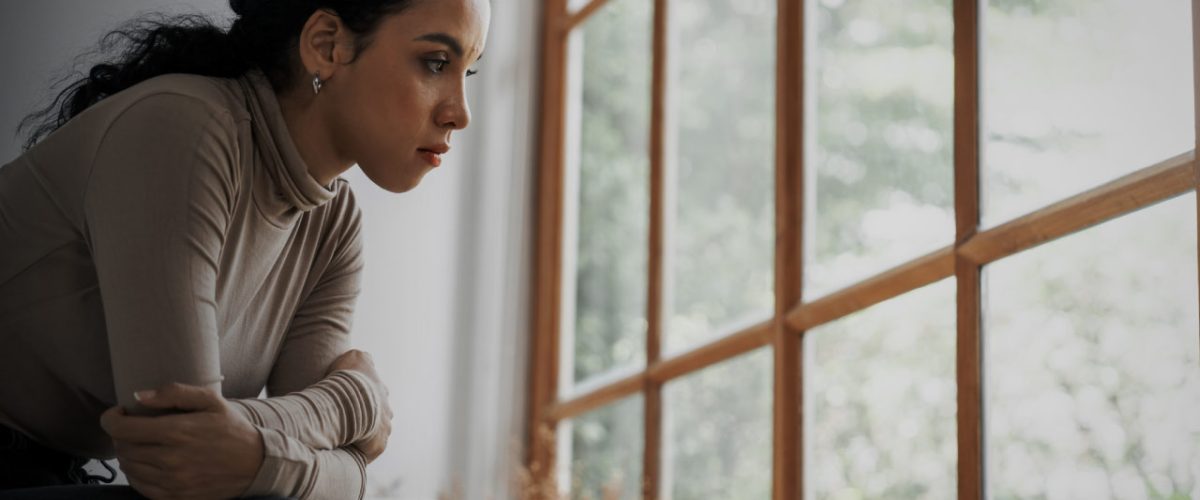 Mulher sentada em frente à janela, com um semblante pensativo e levemente triste.