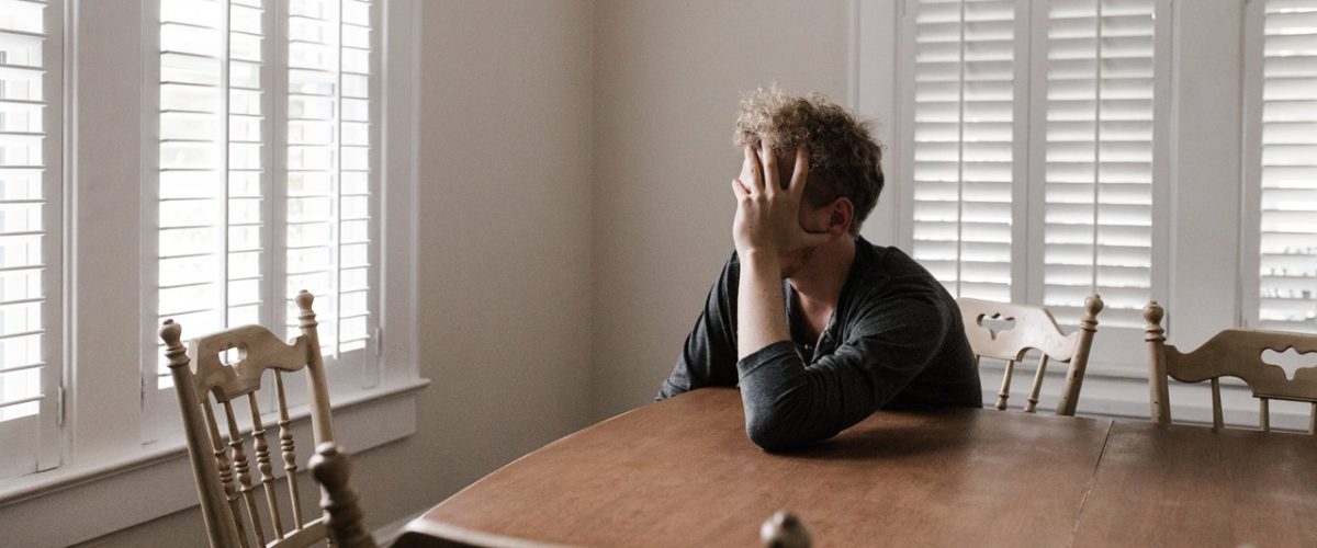 o que é trauma psicológico Homem branco sentado na cadeira, apoiado em uma mesa de madeira, com mão no rosto. Sensação de tristeza.