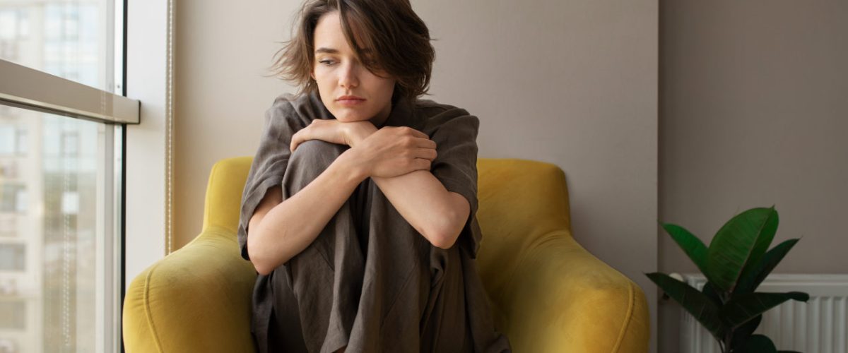 o-que-fazer-quando-a-depressao-passa Mulher com depressão sentada no sofá, ao lado da janela, abraçando os próprios joelhos.