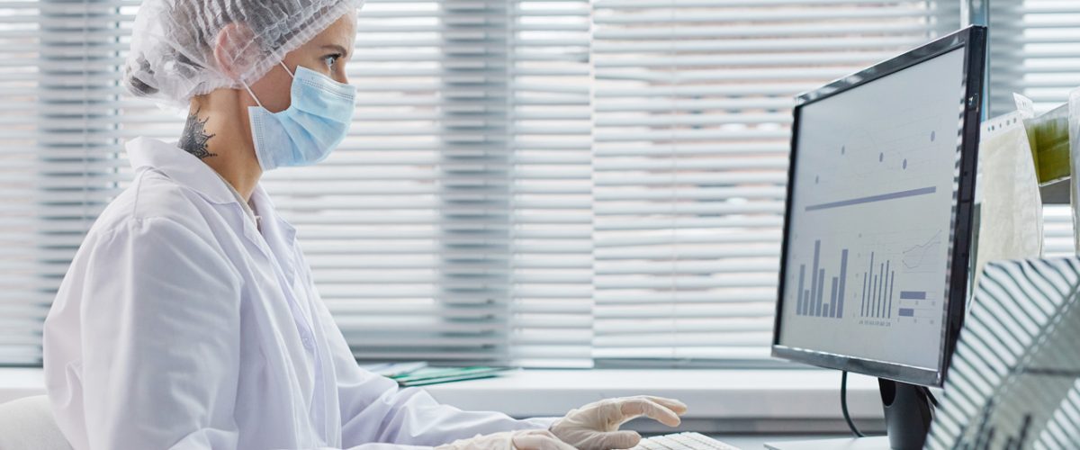 tipos de cetamina Profissional de saúde vestida adequadamente, com toca, bata, máscara e luva, sentada em mesa com computador.